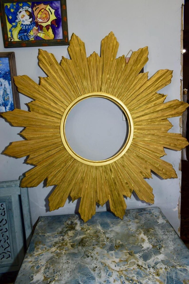 May include: A circular gold sunburst mirror frame featuring a central round opening surrounded by radiating pointed rays. The frame has a textured, metallic gold finish with alternating long and short rays extending outward from the center.