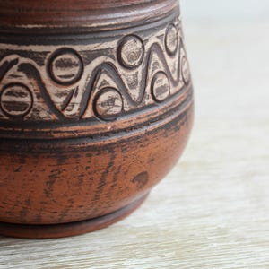 May include: A brown clay vessel featuring a textured surface with a horizontal band of incised geometric patterns. The design consists of repeating circles and zigzag lines filled with a light-colored pigment, contrasting against the dark terracotta clay.