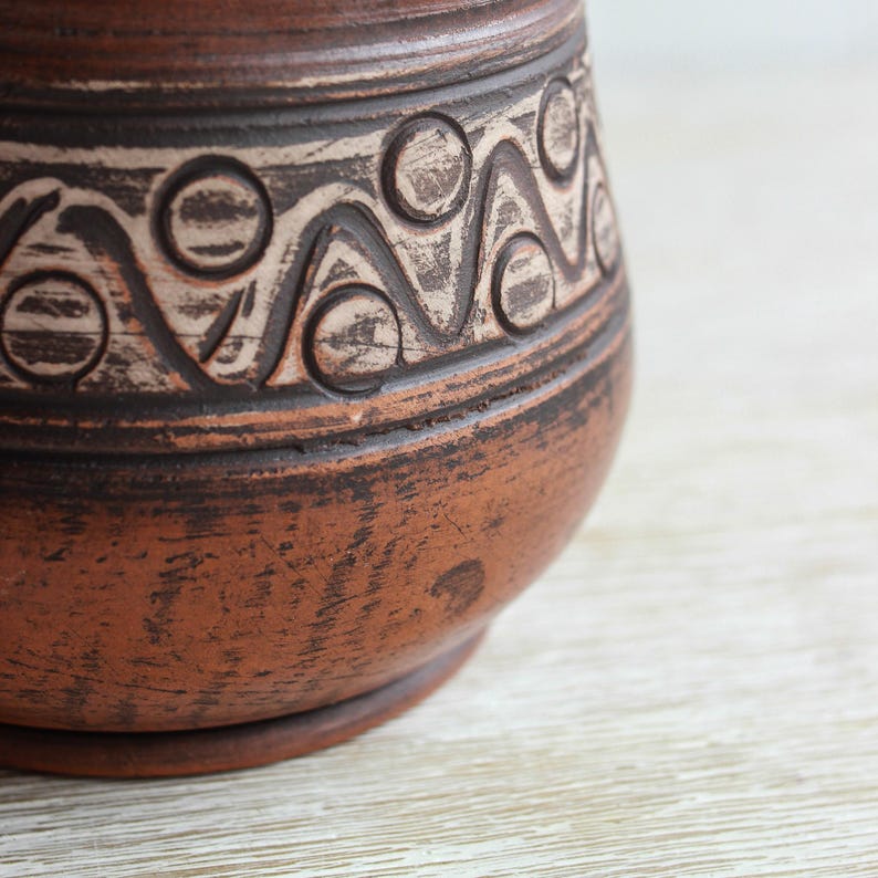 May include: A brown clay vessel featuring a textured surface with a horizontal band of incised geometric patterns. The design consists of repeating circles and zigzag lines filled with a light-colored pigment, contrasting against the dark terracotta clay.