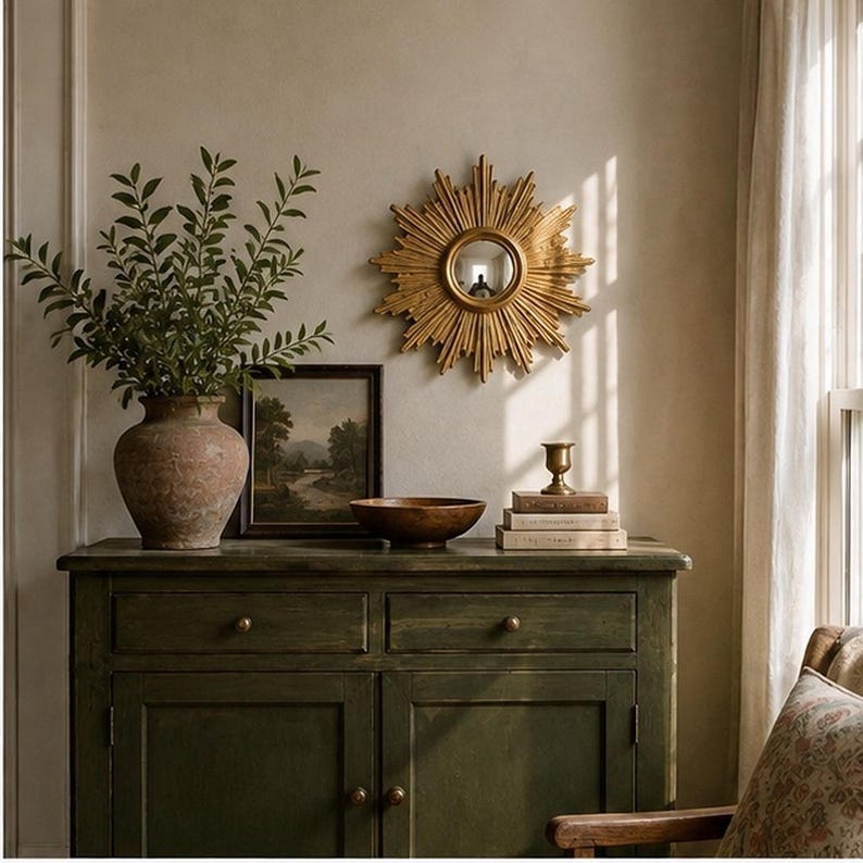 May include: A dark green wooden sideboard with two drawers and two doors. On top sits a large terracotta vase with green foliage, a framed landscape painting, a wooden bowl, a stack of three books, and a small brass candle holder.