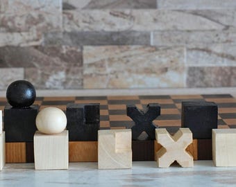 May include: A wooden chessboard with black and white squares. There are wooden pieces on the board, including a black ball, a white ball, and various geometric shapes. The pieces are made of wood and painted black and white.