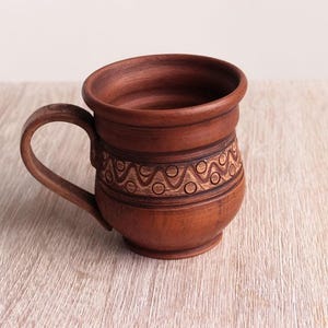 May include: A brown ceramic mug with a decorative pattern around the middle. The mug has a wide, rounded base and a handle.