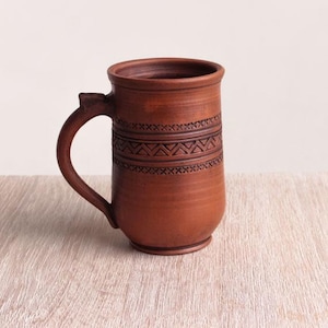 Peut inclure: Une tasse en céramique brune avec une anse et un motif décoratif autour du milieu. La tasse est posée sur une surface en bois brun clair.
