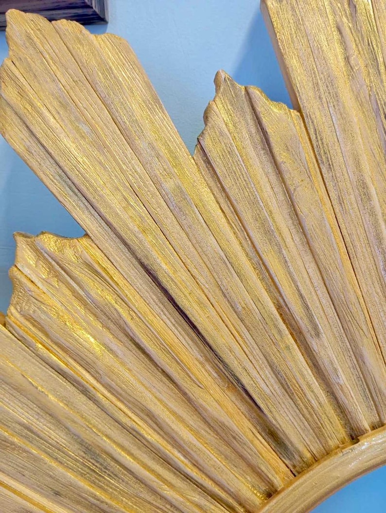 May include: A decorative wall piece featuring a sunburst design with multiple radiating wooden slats. The surface is finished in a metallic gold paint with visible wood grain texture and subtle dark highlights between the individual slats.