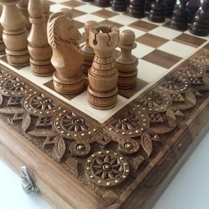 May include: A wooden chess set with intricately carved pieces and a decorative wooden board. The board has a light brown and cream checkered pattern and a carved wooden border with gold accents.