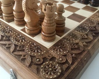 May include: A wooden chess set with intricately carved pieces and a decorative wooden board. The board has a light brown and cream checkered pattern and a carved wooden border with gold accents.