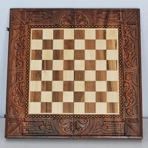 Könnte beinhalten: Ein hölzernes Schachbrett mit geschnitztem Rand. Die Spielfläche ist ein Schachbrettmuster aus hellen und dunklen Quadraten. Der Rand ist aufwendig mit floralen und geometrischen Mustern verziert. Das Schachbrett hat eine satte braune Farbe.