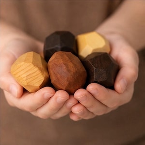 May include: A collection of five small, faceted wooden stones in varying shades of brown, ranging from light tan to dark espresso. Each piece features a smooth, polished surface with visible wood grain patterns and distinct geometric angles.