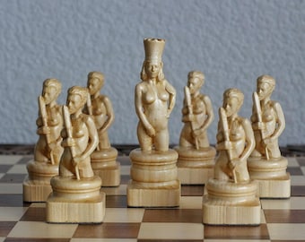 May include: A chess set featuring carved wooden pieces. The pieces are nude female figures, with the queen wearing a tall crown. The chess board has a brown and tan checkered pattern.
