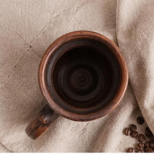 May include: A top-down view of a dark brown, round ceramic mug with a thick, textured rim and a sturdy handle. The interior of the mug is deep and smooth, showing the natural clay finish. The mug is positioned next to scattered coffee beans.