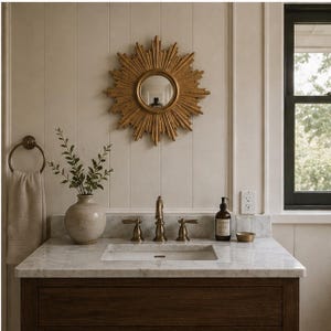 May include: A gold sunburst mirror with a circular center glass, mounted on a vertical paneled wall above a marble countertop vanity. The vanity features a white rectangular sink, a bronze faucet, a ceramic vase with greenery, and a soap dispenser.