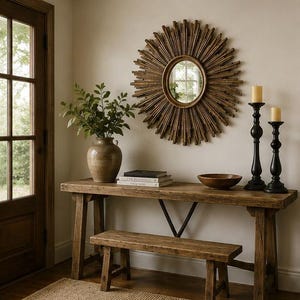 May include: A round sunburst mirror with a dark wood frame hangs above a long wooden console table. The table holds a ceramic vase with green leaves, two stacked books, a wooden bowl, and two black candle holders with cream-colored candles.