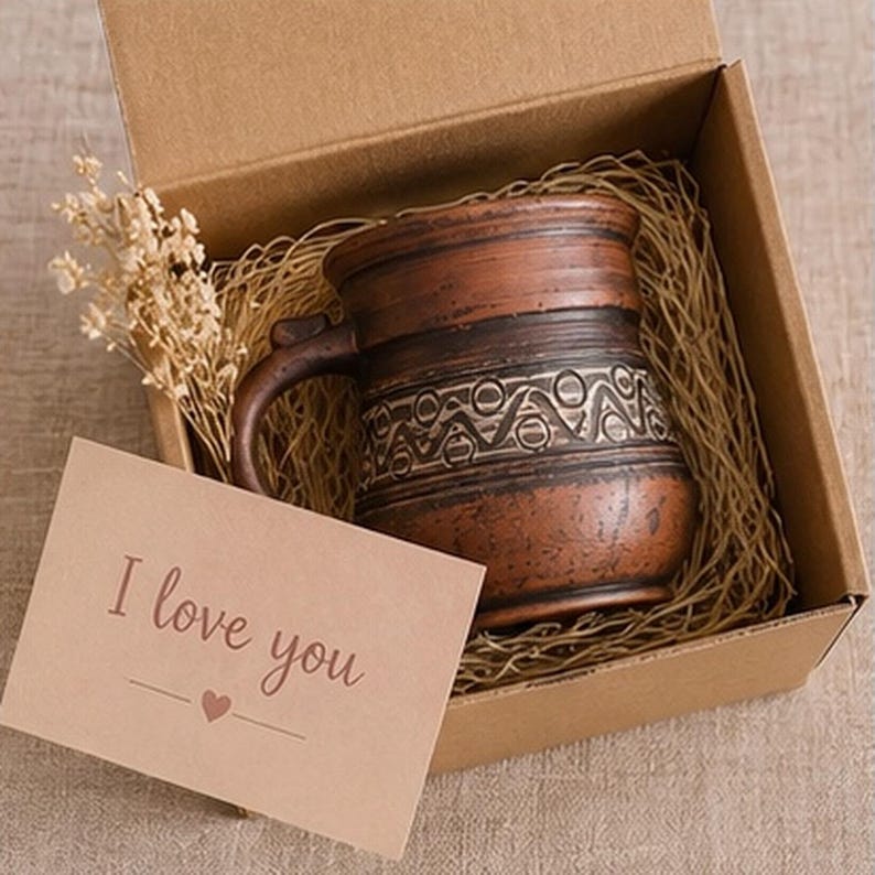 May include: A brown ceramic mug with a textured geometric pattern sits inside a cardboard gift box filled with straw. A small dried flower sprig is tucked beside the mug. A light brown card with the text I love you and a heart icon is included.