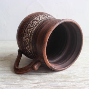 May include: A brown clay mug featuring a wide, flared rim and a curved handle. The exterior surface is decorated with a horizontal band of carved, repeating wave and circle patterns in a lighter, contrasting tone against the dark brown clay.