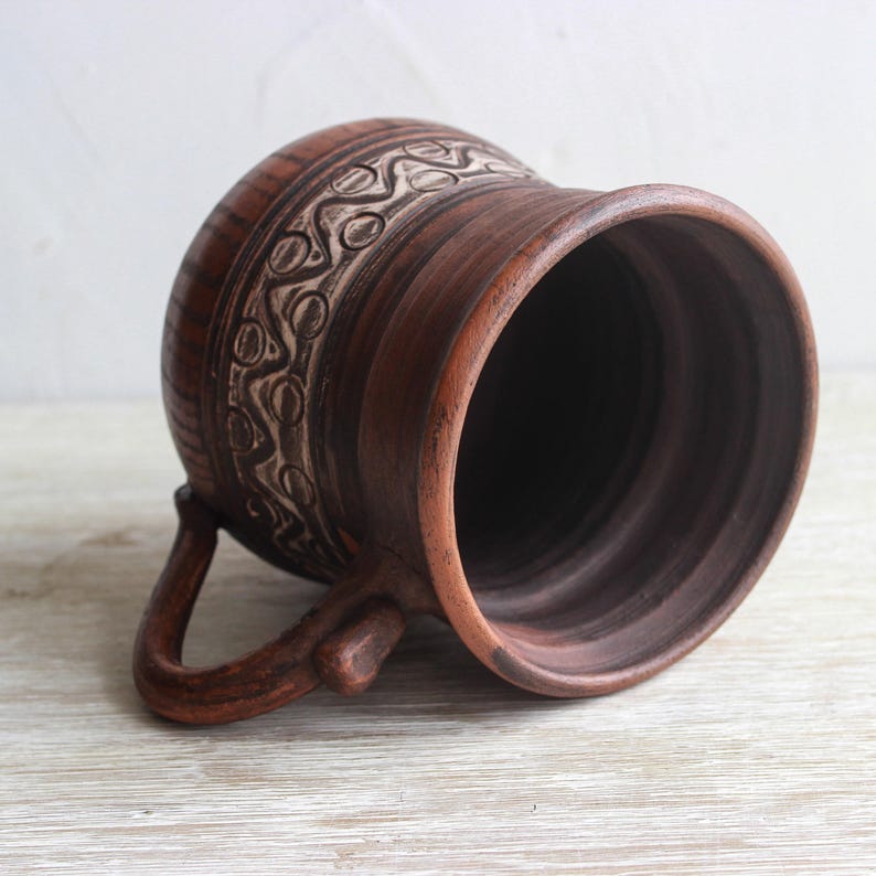 May include: A brown clay mug featuring a wide, flared rim and a curved handle. The exterior surface is decorated with a horizontal band of carved, repeating wave and circle patterns in a lighter, contrasting tone against the dark brown clay.