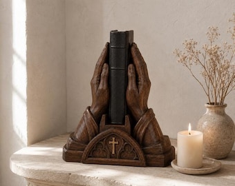 May include: A dark brown resin bookend featuring two sculpted hands pressed together in a prayer position, cradling a black book. The base is decorated with a small gold-colored cross set within an arched, gothic-style architectural frame.