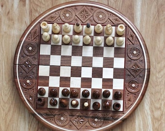 May include: A wooden chess set with a round, intricately carved board. The board is open and shows a game in progress with light and dark pieces on a checkered board.