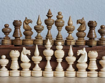 May include: A set of wooden chess pieces in two colours: brown and white. The pieces are carved with intricate details and have a traditional design. The chess pieces are arranged on a wooden chessboard.