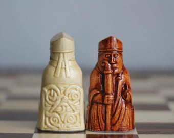 May include: Two chess pieces, a bishop and a king, are on a chessboard. The bishop is brown and has a detailed design. The king is white and has a Celtic knot design.