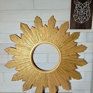 May include: A circular gold sunburst mirror frame featuring a central round opening surrounded by numerous radiating pointed rays of varying lengths. The frame has a textured, metallic gold finish with visible brush strokes throughout the surface.