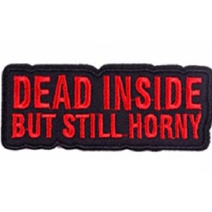 May include: A rectangular black patch with red embroidered text that reads "DEAD INSIDE BUT STILL HORNY". The patch has a black border and is likely designed to be sewn or ironed onto clothing or accessories.