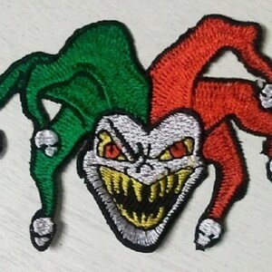 May include: Embroidered patch featuring a menacing jester design. The patch has a green and red jester hat with skull accents. The jester's face is white with yellow teeth and red eyes, creating a scary look.
