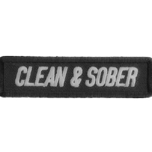 May include: A rectangular black patch with the words "CLEAN & SOBER" embroidered in white. The patch has a textured appearance, and the text is in a bold, sans-serif font. The patch is likely designed to be sewn onto clothing or accessories.