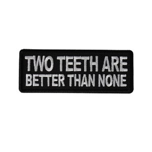 May include: Black rectangular patch with white embroidered text that reads "TWO TEETH ARE BETTER THAN NONE". The patch has a black border and is likely designed to be sewn or ironed onto clothing or accessories.