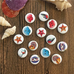 May include: Assortment of 14 round wooden buttons with various sea life designs. The buttons feature images of seashells, starfish, seahorses, and a nautilus shell. The buttons are white with colorful designs.