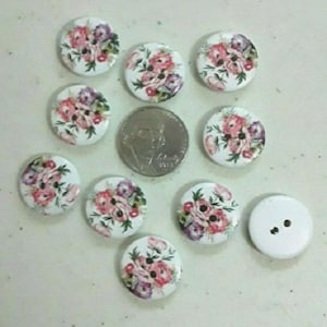 May include: Assortment of ten round wooden buttons with floral designs. The buttons are white with pink and purple flowers. One button is plain white with two holes.