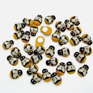 May include: A collection of small, hand-painted bee-shaped embellishments. Each bee features a yellow body with black stripes and white wings. The bees are scattered on a white surface, perfect for craft projects.