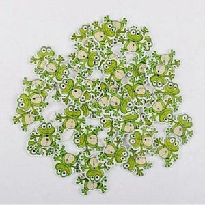 May include: A collection of small, wooden frog-shaped buttons. Each button features a green frog with a white belly and large, cartoonish eyes. The buttons are scattered across a white surface.