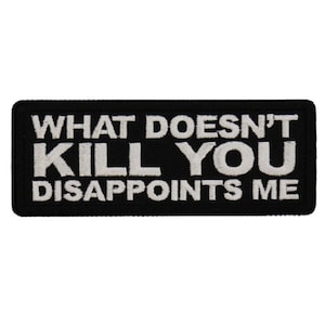May include: Black rectangular patch with white embroidered text that reads "WHAT DOESN'T KILL YOU DISAPPOINTS ME". The patch has a black border and is likely designed to be sewn or ironed onto clothing or accessories.