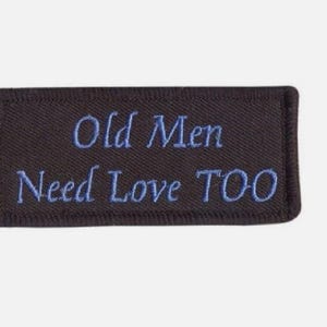 May include: A rectangular black embroidered patch with a stitched border. The text "Old Men Need Love TOO" is embroidered in light blue thread across the center of the patch in a serif font, with the word "TOO" appearing in capital letters.