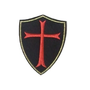May include: A black embroidered patch with a red cross inside a gold shield. The cross has a pointed top and bottom.
