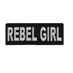 May include: A rectangular black patch with the embroidered text "REBEL GIRL" in white block letters. The patch is likely designed to be sewn or ironed onto clothing or accessories.