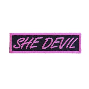 May include: A black rectangular patch with pink stitching around the edges. The text "SHE DEVIL" is embroidered in pink.