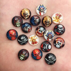 May include: Assortment of colorful round skull and pirate themed cabochons. The cabochons are approximately 1 inch in diameter and feature various designs, including skulls with crossbones, pirates with swords, and skulls with crowns.