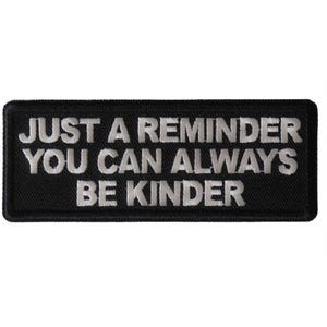 May include: A rectangular black embroidered patch with a stitched border. The text "JUST A REMINDER YOU CAN ALWAYS BE KINDER" is displayed in white, capitalized, sans-serif lettering across three lines, centered on the black background.