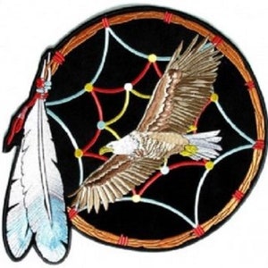 May include: A black and brown embroidered patch featuring a dreamcatcher with a white eagle flying through it. The dreamcatcher has red, white, and blue accents. Two white feathers hang from the bottom of the dreamcatcher.