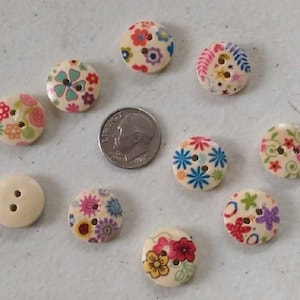 May include: Ten round, light-colored wooden buttons featuring various colorful floral patterns, each with two holes. A silver-colored United States dime is positioned in the center for scale, measuring approximately 0.7 inches in diameter.