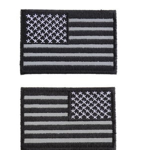 May include: Two black and white American flag patches. The flags are embroidered with white thread on a black background. The patches are rectangular and have a stitched border.