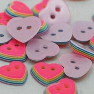 May include: A collection of heart-shaped buttons in shades of pink and rainbow colors. Some buttons are solid pink, while others have a layered rainbow effect. Each button has two holes for sewing.