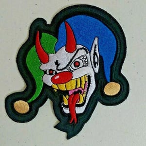 May include: An embroidered patch featuring a jester-like character with a white face, red nose, and red horns. The character wears a green and blue cap with gold bells. It has yellow teeth, a red tongue, and is outlined in a thick black border.