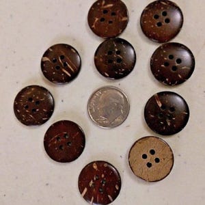 May include: Nine circular dark brown buttons with four holes each, featuring natural wood grain patterns and textures. A silver-colored United States dime is positioned in the center of the arrangement to provide a scale reference for the buttons.