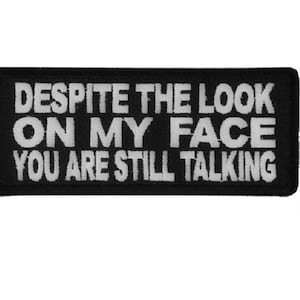 May include: A rectangular black embroidered patch with a white border. The text in white capital letters reads: DESPITE THE LOOK ON MY FACE YOU ARE STILL TALKING. The patch has a clean, stitched edge and a high-contrast design for clear readability.