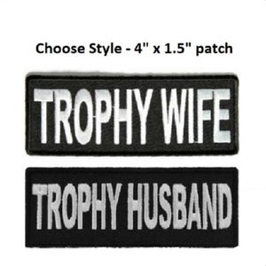 May include: Two rectangular black embroidered patches with white text. The top patch reads TROPHY WIFE and the bottom patch reads TROPHY HUSBAND. Each patch measures 4 inches by 1.5 inches as indicated by the text Choose Style - 4 x 1.5 patch.