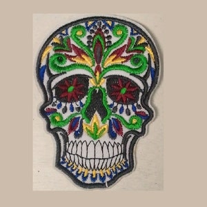 May include: A colorful embroidered patch featuring a white sugar skull with green, red, blue, and yellow floral designs. The skull has a black nose and black eyes with red and white flower accents.
