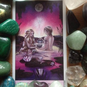 Puede incluir: Ilustraci&oacute;n de carta de tarot que representa dos figuras en el agua, rodeadas de nen&uacute;fares, bajo un cielo iluminado por la luna. La carta est&aacute; enmarcada por varias piedras preciosas de colores, incluyendo malaquita verde y cuarzo transparente.