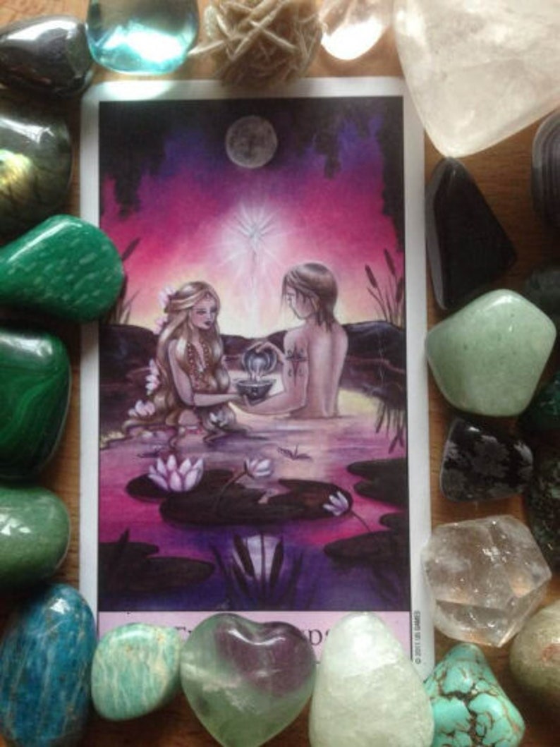 Puede incluir: Ilustraci&oacute;n de carta de tarot que representa dos figuras en el agua, rodeadas de nen&uacute;fares, bajo un cielo iluminado por la luna. La carta est&aacute; enmarcada por varias piedras preciosas de colores, incluyendo malaquita verde y cuarzo transparente.